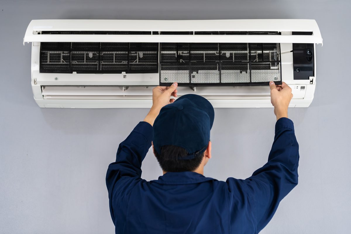 A technician in a navy cap and overalls, seen from behind, lifting the front panel of a wall-mounted heat pump to reach its filters