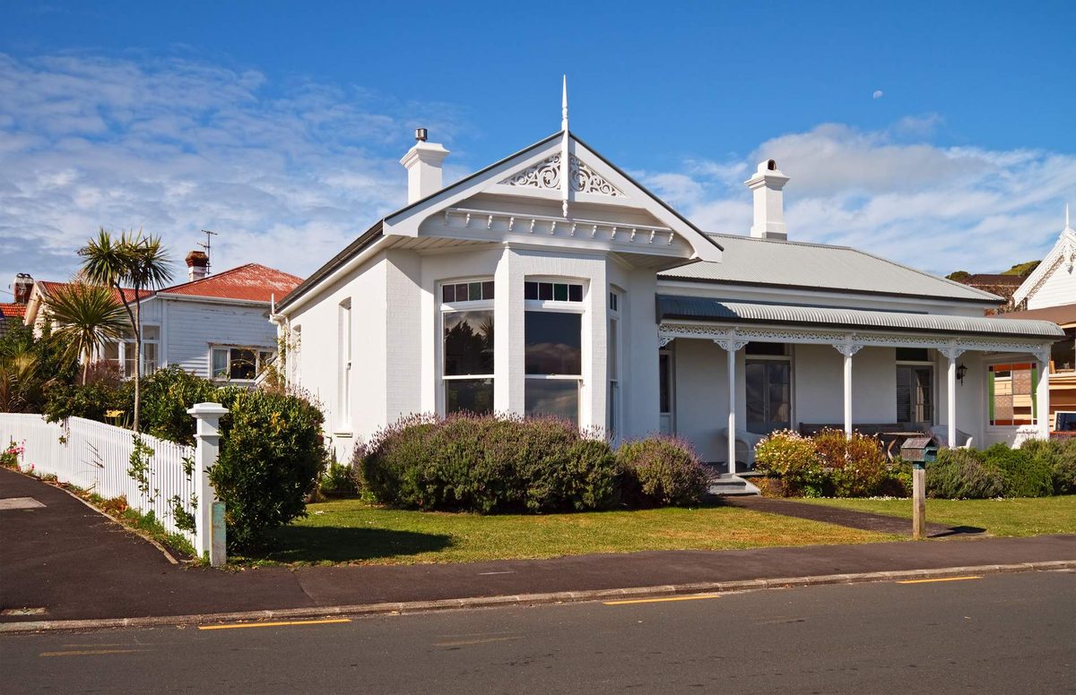 A single-storey white villa with a bay window, carved gable trim, two chimneys and a front verandah, behind a picket fence and garden